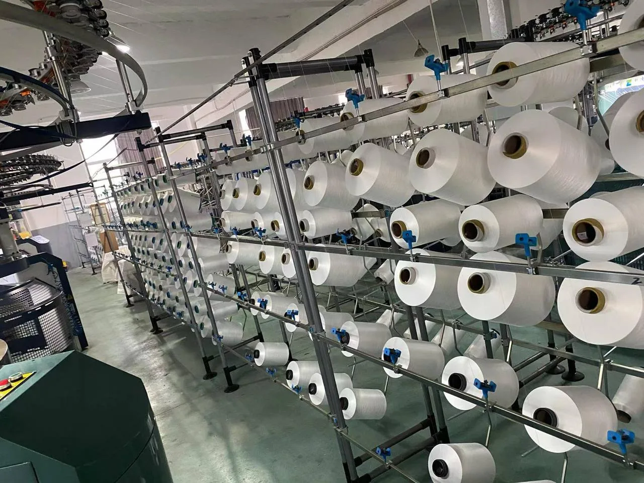 Reihen weißer Polyester-Garnspulen im Gatter neben einer Strickmaschine